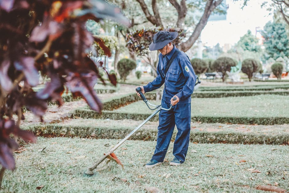 A gardener wearing a blue uniform, hat, and glasses is using a handheld grass trimmer to cut the grass in a well-maintained park. The lawn features neatly trimmed grass and cleanly edged garden beds b